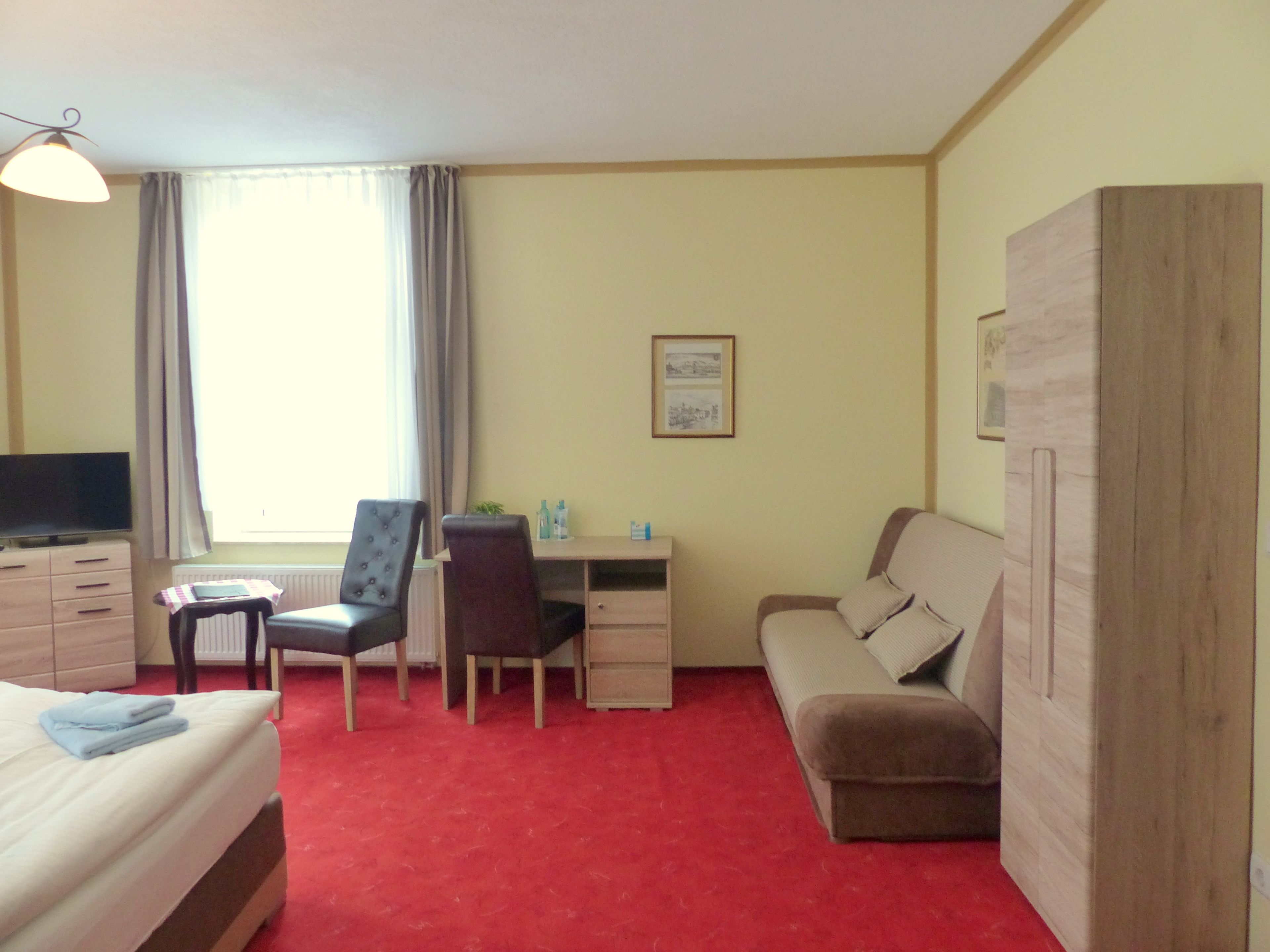 A hotel room with a bed, a desk, a chair, a sofa, a wardrobe, a television, and a window with curtains.
