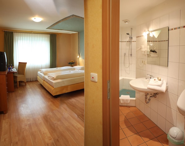 Habitación de hotel con cama, escritorio con silla, televisor y baño con lavabo, espejo y ducha.