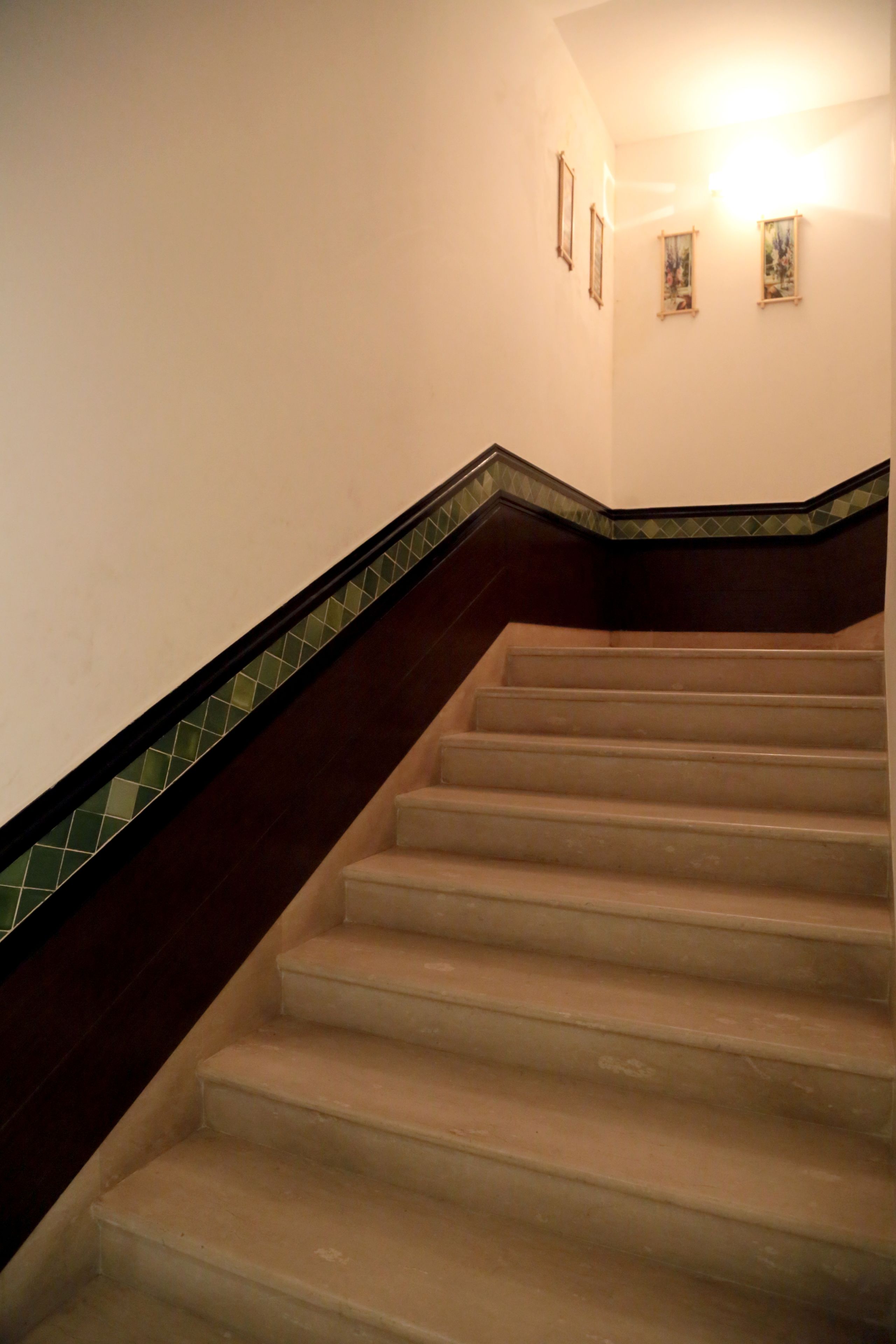 Una escalera con barandal de madera oscura y escalones color beige, que conduce a un rellano con una lámpara.