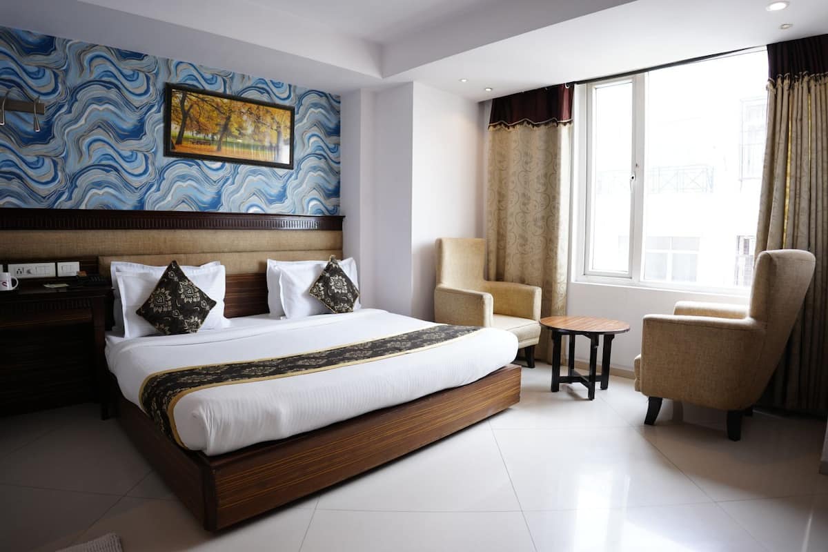 A hotel room with a large bed, a chair, a small table, a nightstand, and a window with curtains.