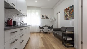 A modern kitchen with a kettle, toaster, and a coffee machine. There is a dining area with a table and chairs, and a sofa with a bookshelf. - Italianway   - Segrino 6 (Milan)