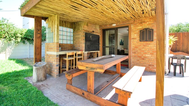 An outdoor patio with a wooden table and benches, a brick fireplace, and a covered area with a slatted roof. - Coral Sands Brickstone (Cape Town)