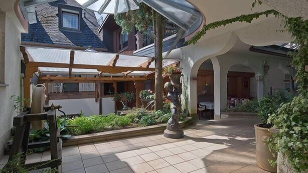 A covered patio area with a pergola, a statue, and a view of a house through a glass ceiling. - Hotel Restaurant Schütz (Trier)