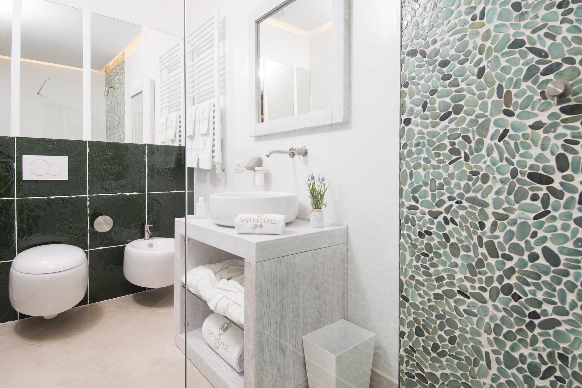 Un baño moderno con un lavamanos blanco, un inodoro y una ducha. Hay un espejo sobre el lavamanos y una pared con mosaicos.