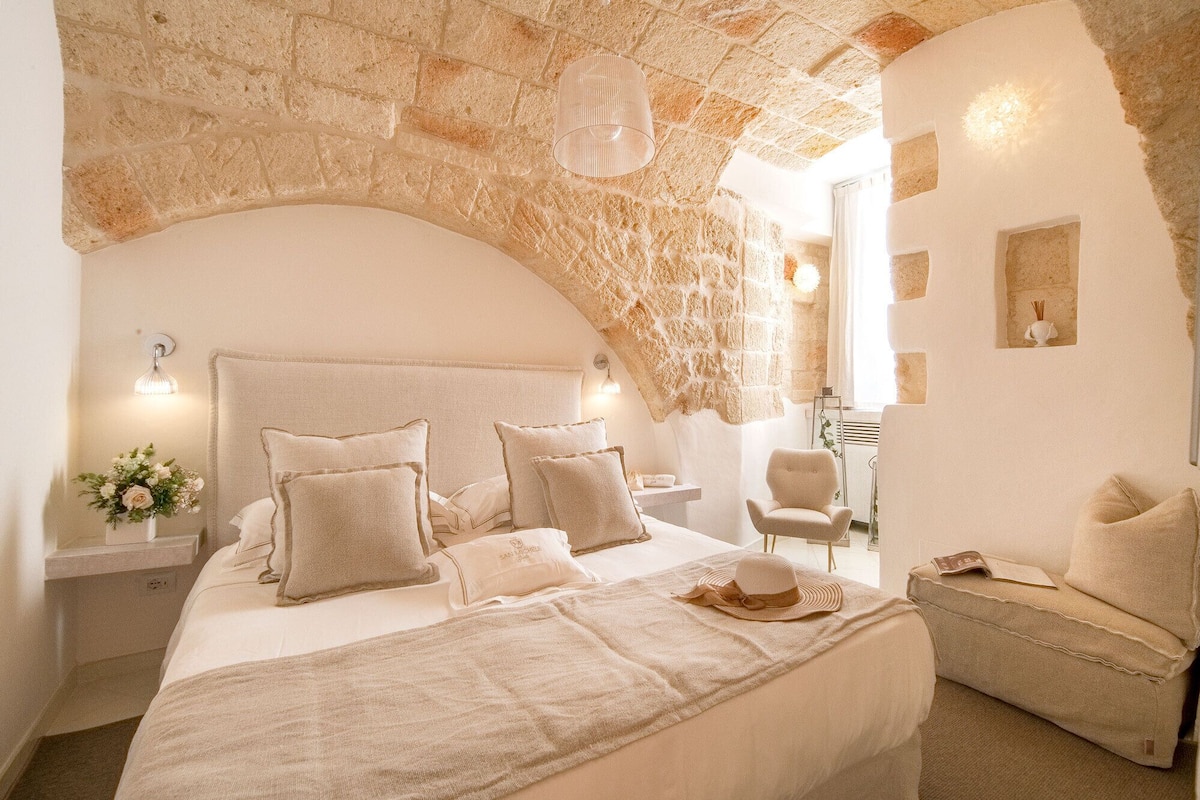 Un dormitorio con un arco de piedra, una cama con muchos almohadones, una mesita de noche con una lámpara y una silla.