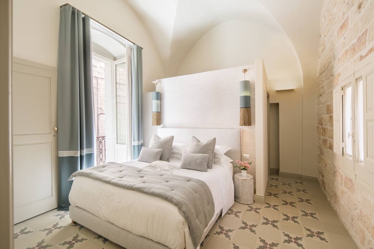 Un dormitorio con una cama grande, cabecera, mesita de noche y ventana con cortinas.