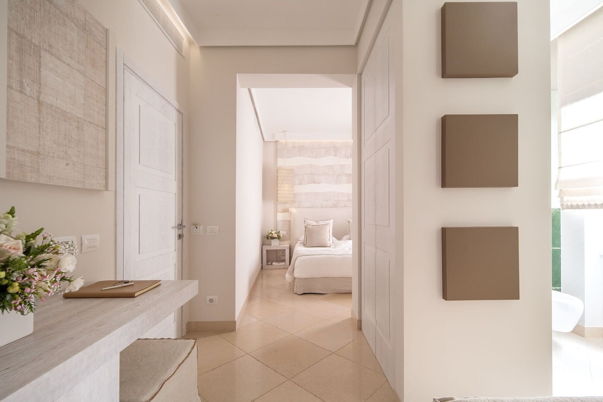 Un pasillo que conduce a un dormitorio con una cama, una mesita de noche y una lámpara fijada en la pared.