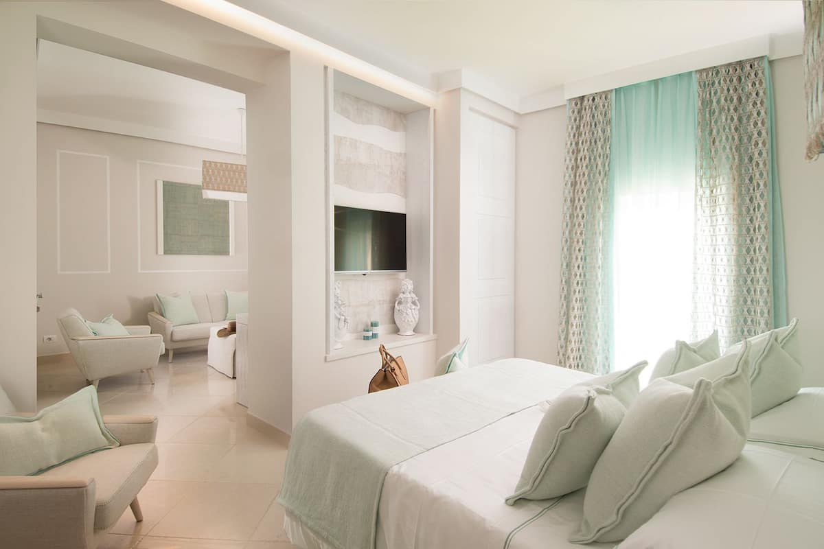 Una habitación de hotel moderna con una cama grande, un área de estar con sofá y sillones, un televisor montado en la pared y un ventanal con cortinas estampadas.