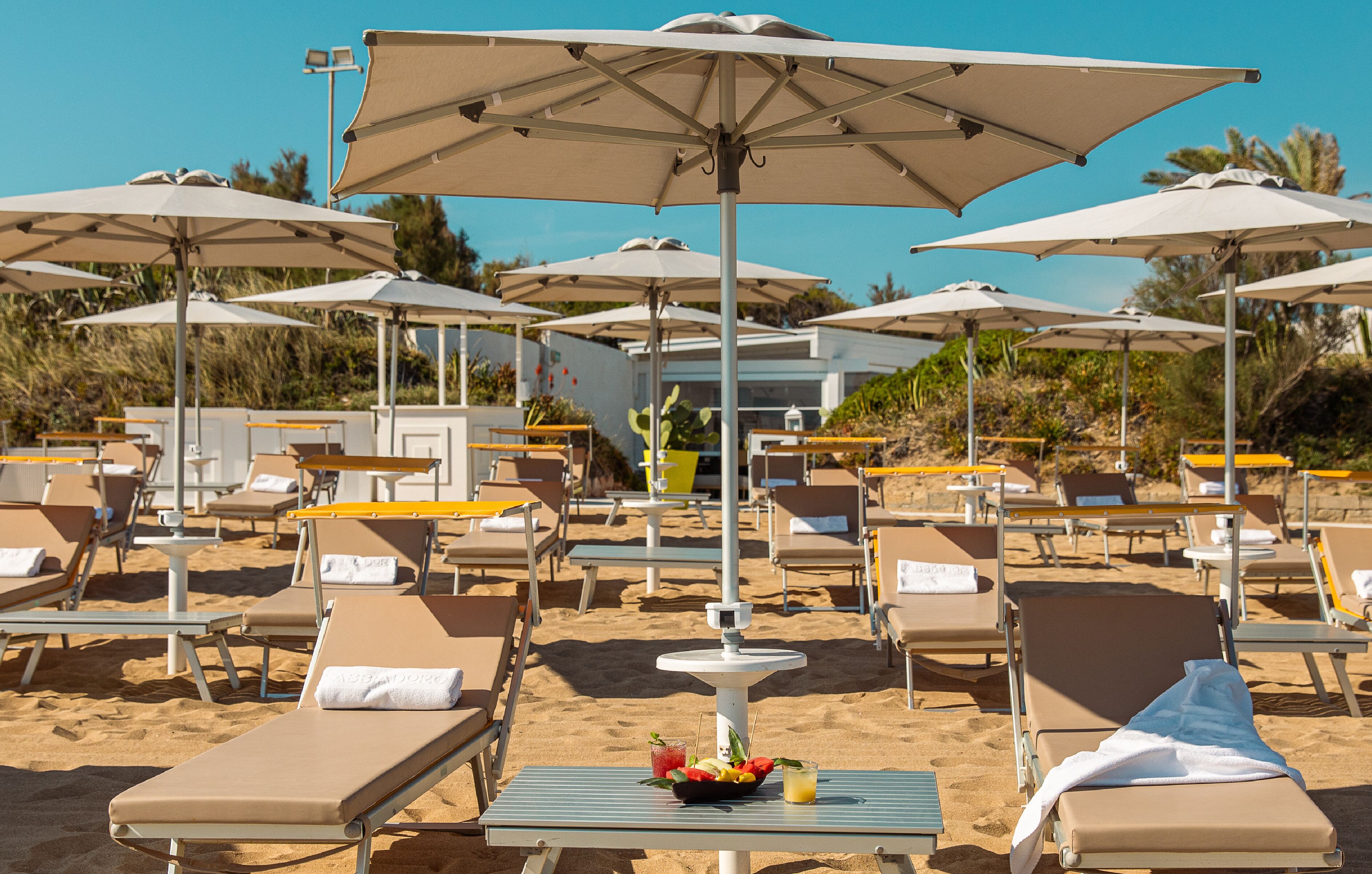 Een loungeruimte aan het strand met ligbedden, parasols en een tafel met een schaal fruit en een drankje.