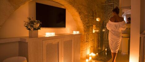 A spa area with a stone wall, a mounted TV, a reception desk with a flower vase, and a person in a robe near a sink.