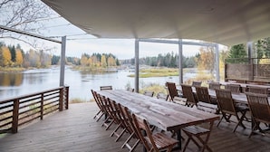 Et overdækket spiseområde med træborde og stole, med udsigt over en flod og efterårsfarver.