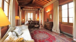 A room with a bed, a wooden ceiling, a dining table with chairs, a dresser, and a large window with curtains. - Villa Il Colle (Bagno a Ripoli)