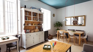 A bright living room with a wooden table, chairs, a bookshelf, and a large mirror. - La Casa Morada - Hostel (Cádiz)