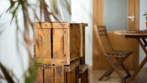 A wooden crate with a picture frame leaning against it, a wooden chair, and a dining table in a room with a door and a plant in the background. - La Casa Morada - Hostel (Cádiz)