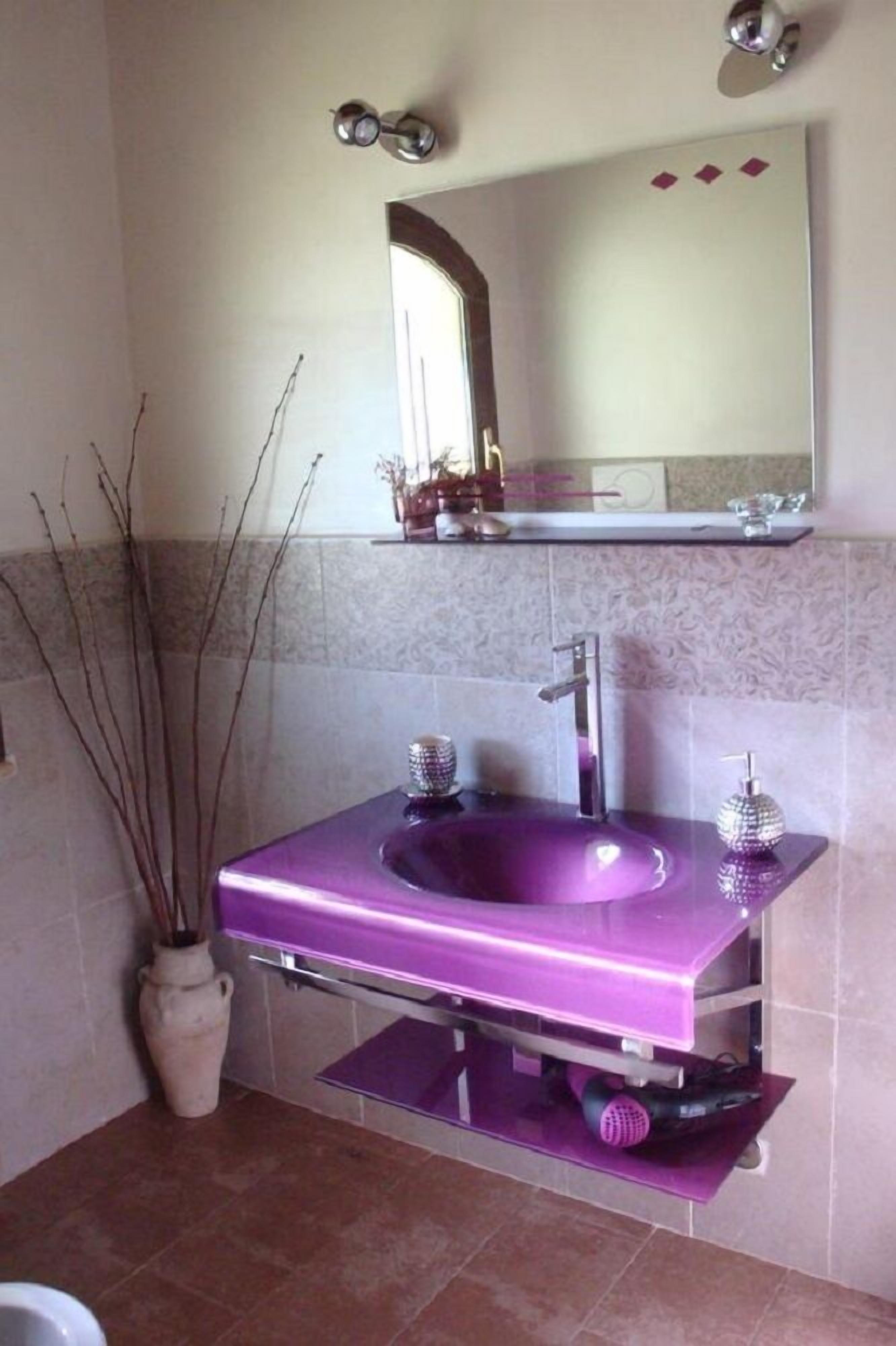 Une salle de bain avec un lavabo violet, un miroir au-dessus et un vase avec des branches sur la gauche.