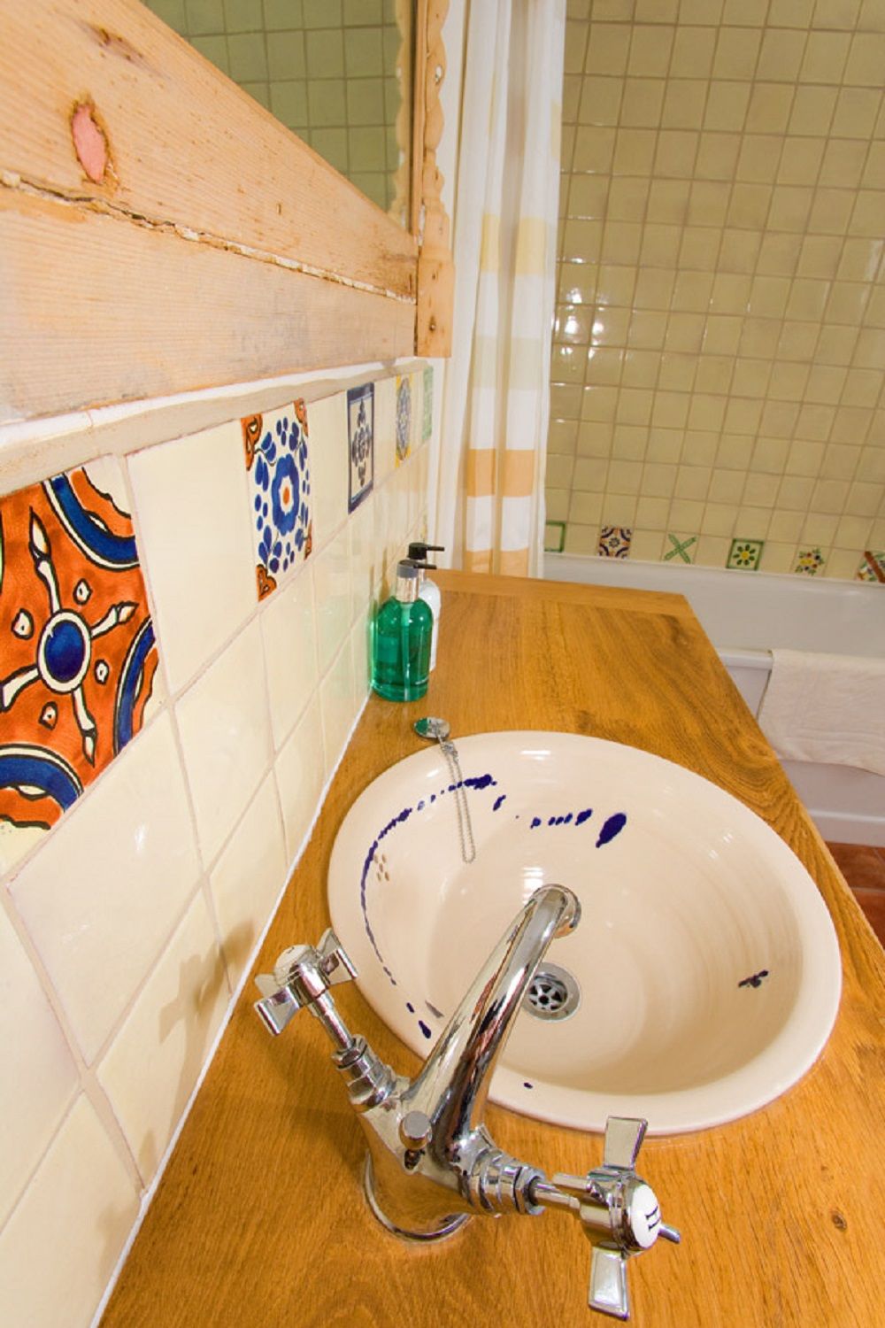 Un baño con tocador de madera, un lavabo blanco con manchas azuladas de agua, un grifo cromado y una botella de jabón líquido verde.