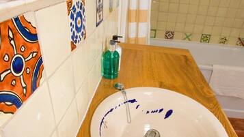 Un baño con tocador de madera, un lavabo blanco con manchas azuladas de agua, un grifo cromado y una botella de jabón líquido verde.