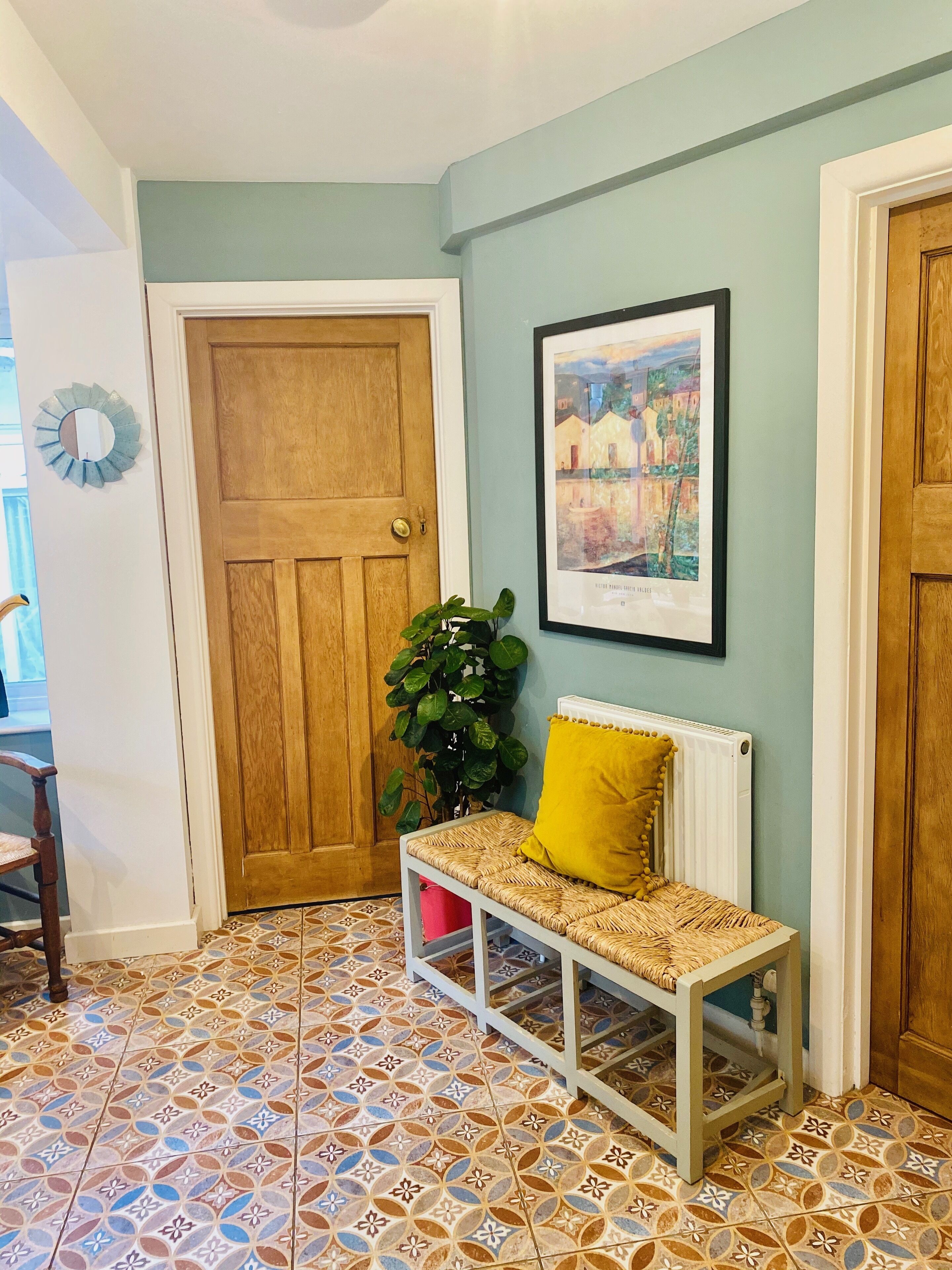 A hallway with a wooden door, a bench with a yellow cushion, a potted plant, and a framed picture on the wall.