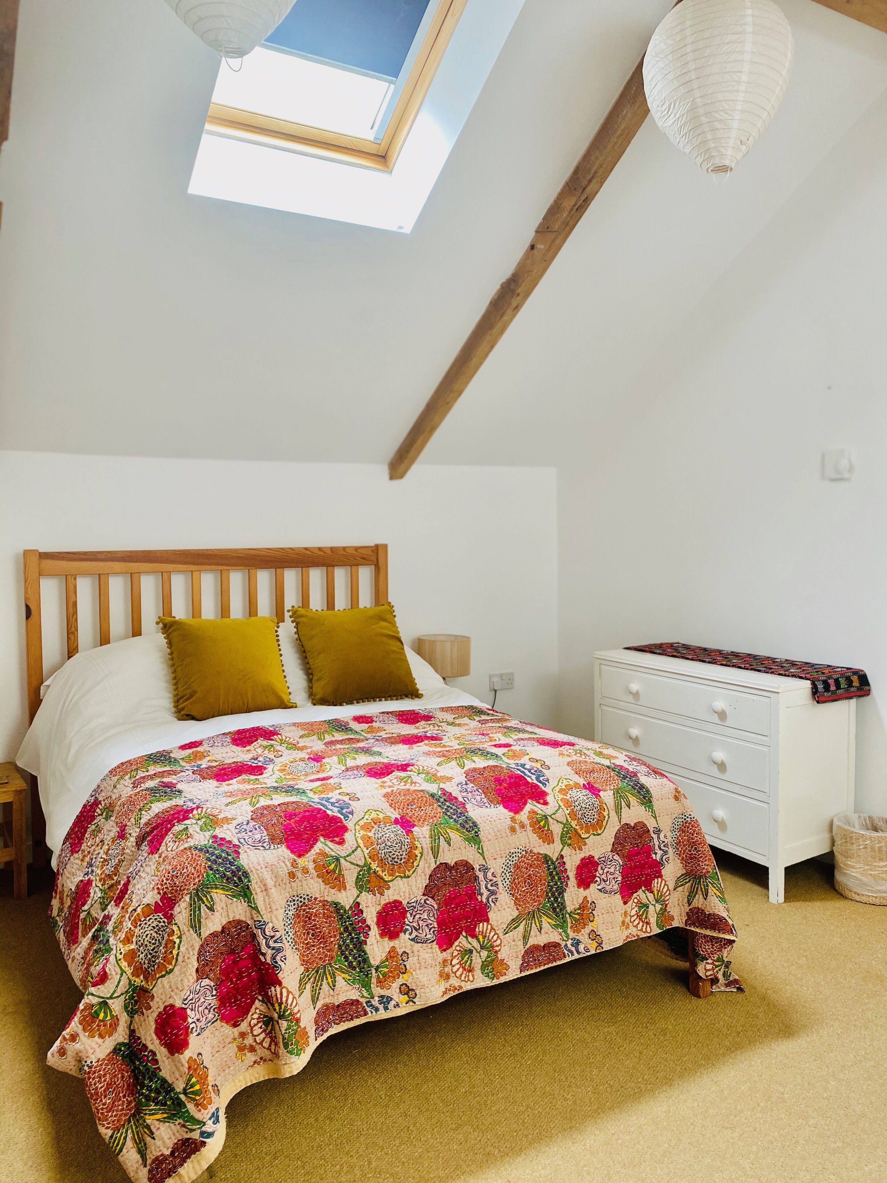 A bedroom with a wooden bed, a colorful floral bedspread, a white nightstand, a white dresser, and a skylight above.