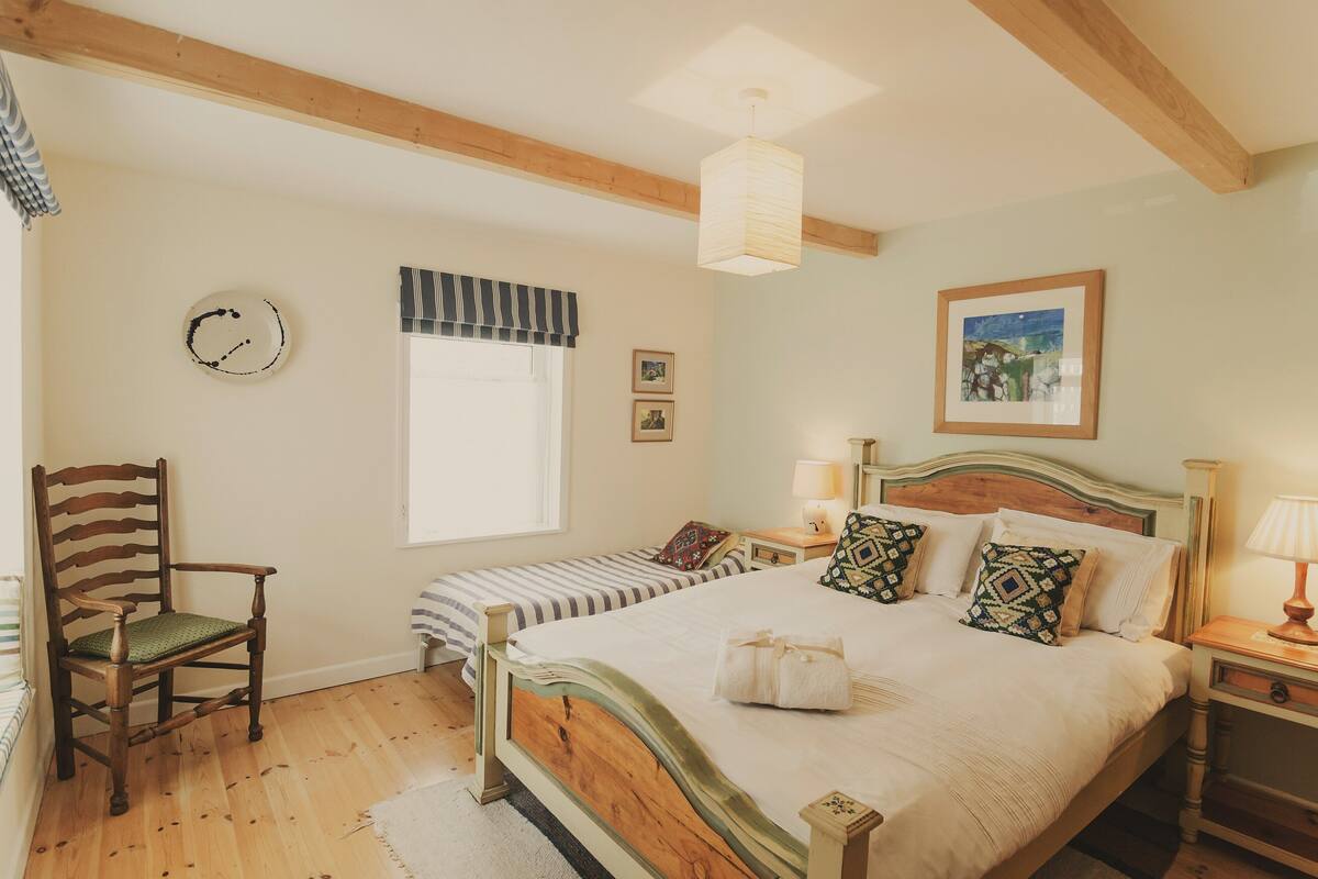 A bedroom with a wooden bed, a chair, a bedside table with a lamp, a window with a striped valance, and a wall clock.