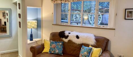 Ein braunes Ledersofa mit floralen Kissen, ein Fenster mit dekorativer Vorhangstange und Blick auf eine große Radkonstruktion draußen.