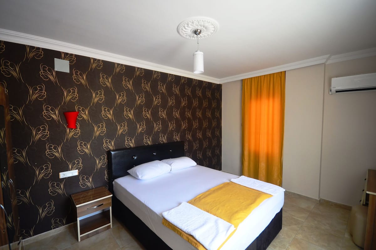 Quarto de hotel com cama, mesa de cabeceira, vaso vermelho, luminária de teto branca e janela com cortinas amarelas.