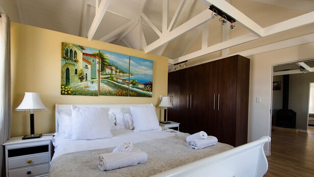 A neatly made bed with white linens and towels, a painting of a coastal scene on the wall, and a wooden wardrobe. - The Bicycle Cottage (Hermanus)