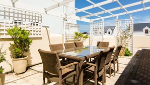 A covered patio with a glass table and wicker chairs, surrounded by potted plants. - The Bicycle Cottage (Hermanus)