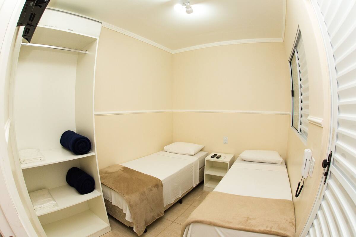 Una habitación de hotel pequeña y sencilla, con cama, mesita de noche, armario y ventana con persianas.