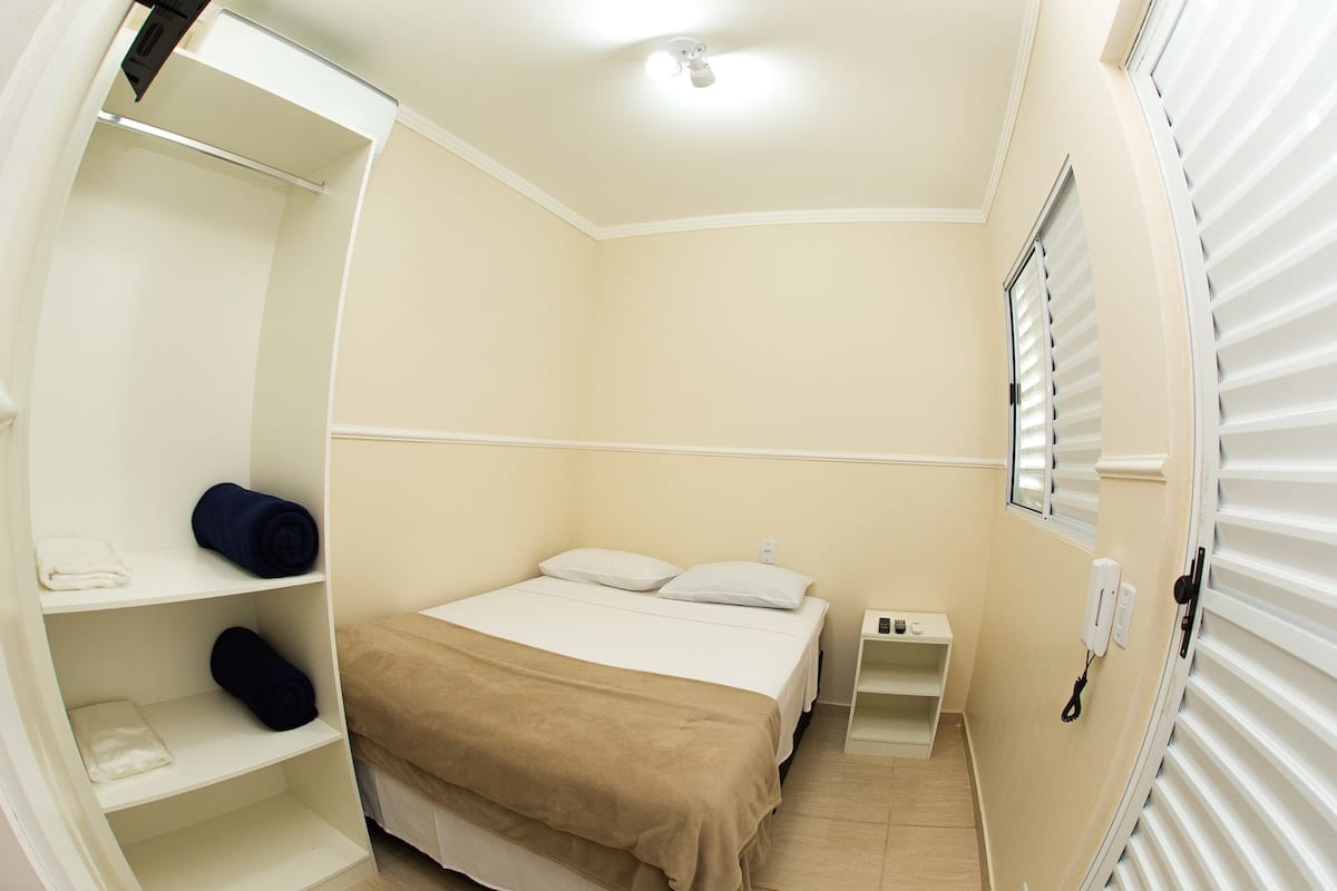 Un dormitorio pequeño y bien iluminado con una cama individual, una mesita de noche, un armario con estantes y una ventana con persianas.