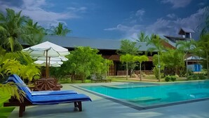 A pool area with lounge chairs and umbrellas, surrounded by palm trees and a building with a sloped roof. - Silver Crown Nature Resort (Anuradhapura)