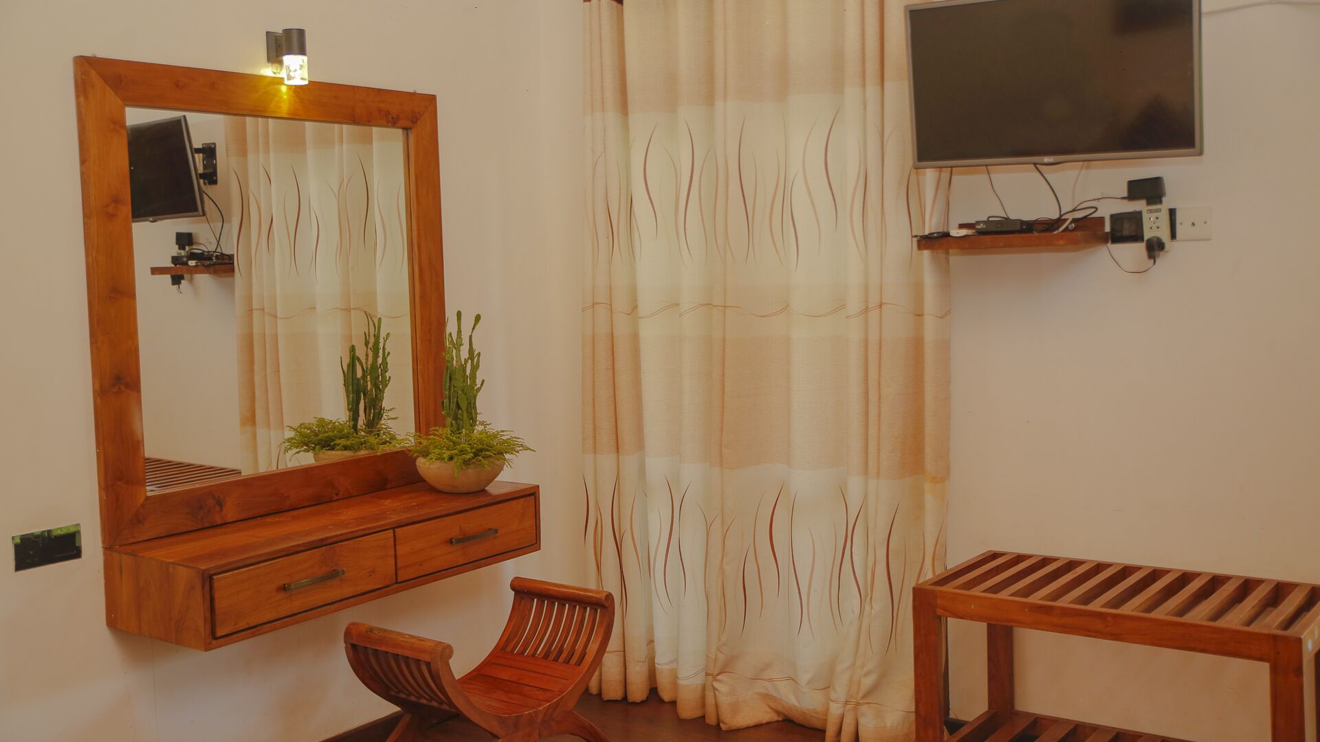 A room with a wooden cabinet, a chair, a small table, a television mounted on the wall, and a curtain with a wavy pattern.