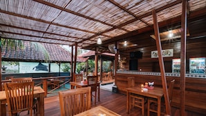 Een openluchtrestaurant met houten tafels en stoelen, een overdekt gedeelte met rieten dak en uitzicht op het omliggende landschap.