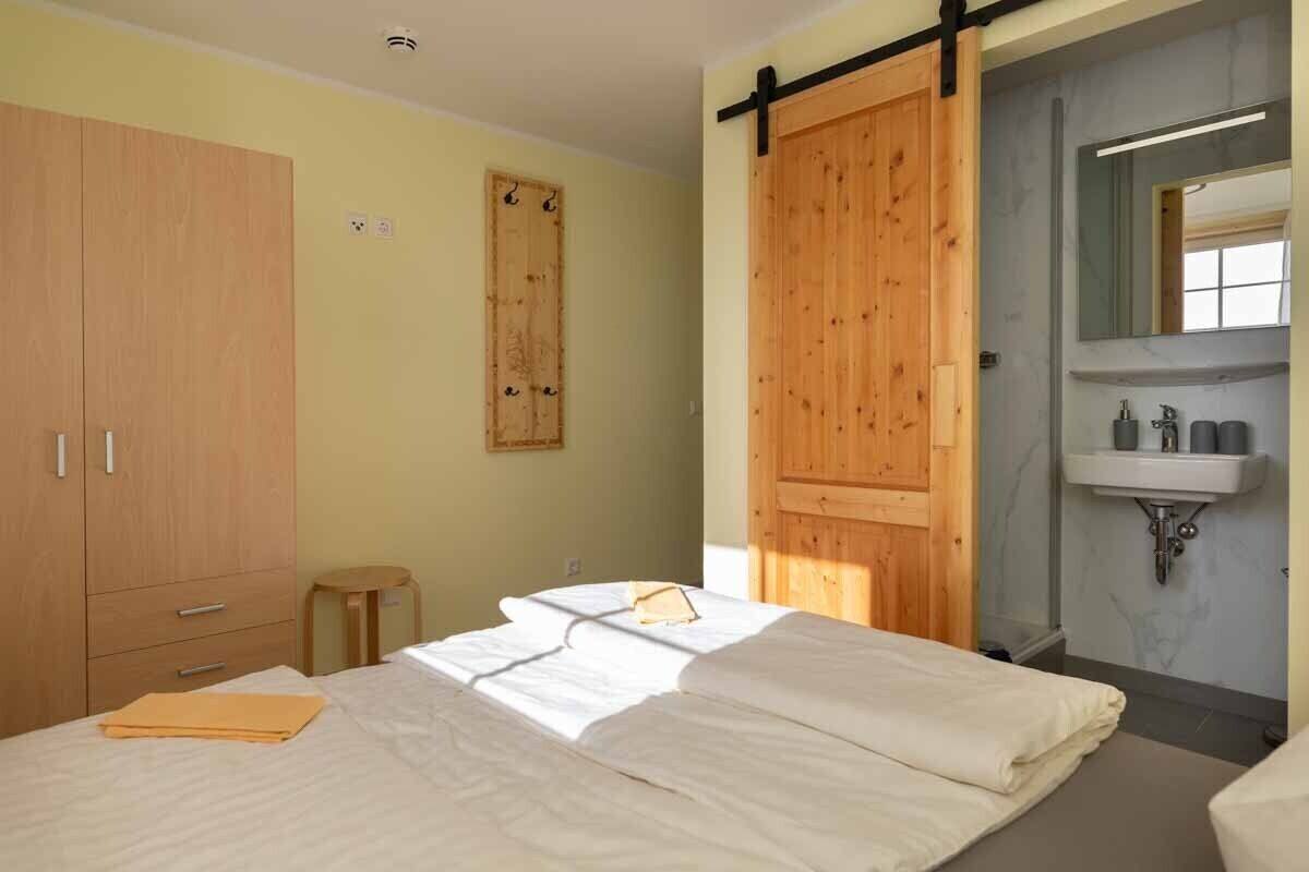 A bedroom with a bed, wooden wardrobe, a small stool, a wooden door, and a bathroom with a sink and mirror.