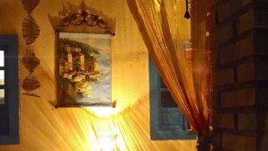Une pièce éclairée d’une lumière chaleureuse, avec un tableau, une table sur laquelle est posé un vase et une fenêtre ornée de rideaux.