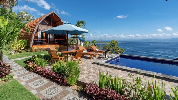 Una villa frente a la playa con piscina, sillas de descanso y una estructura de techo de paja.