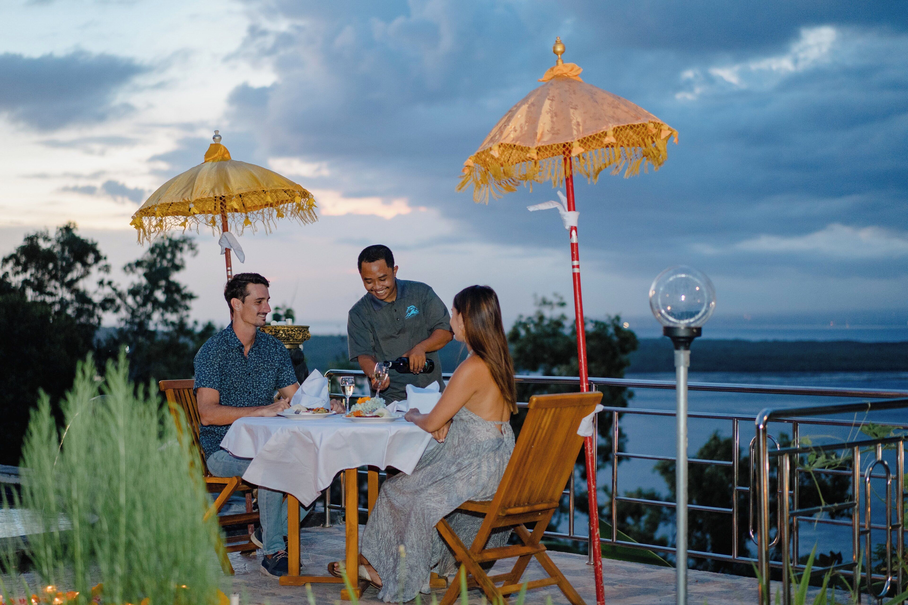 A table set outdoors with a view of the water, surrounded by wooden chairs and yellow umbrellas.