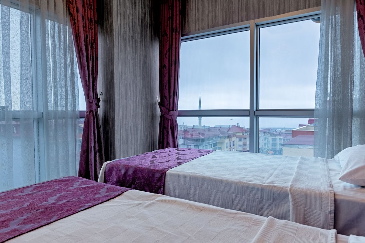 Habitación de hotel con dos camas, un ventanal con cortinas y vistas a un paisaje urbano.