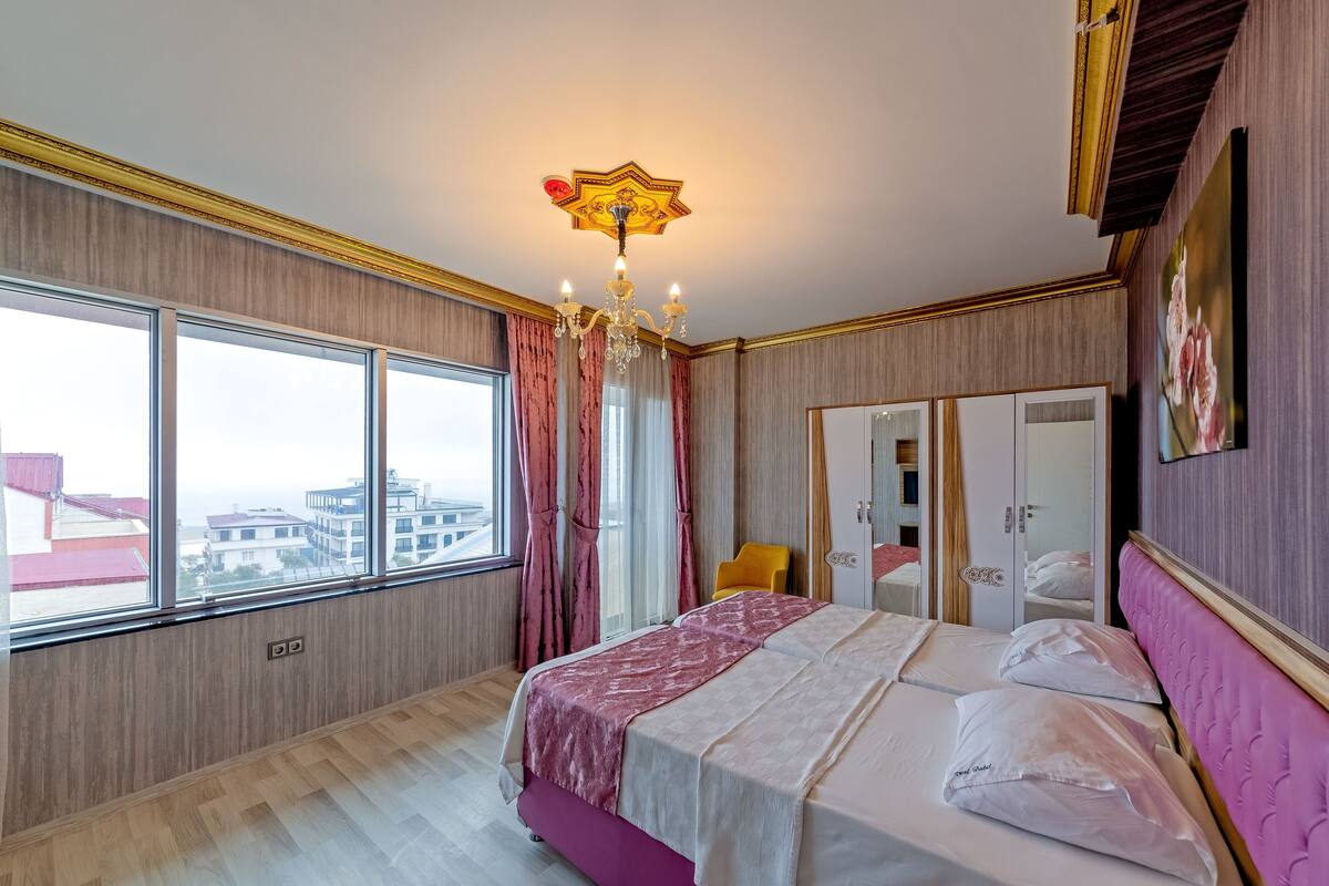 Un dormitorio con una cama grande, un candelabro, un armario y vistas a edificios a través de amplios ventanales.