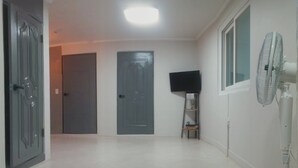 Un pasillo con suelo llano, una puerta, un televisor sobre un mueble bajo y un ventilador de pie.