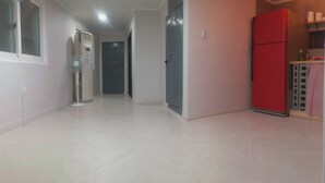 Un pasillo con piso de baldosas blancas, un ventilador de pie, un refrigerador y una puerta que da a otra habitación.
