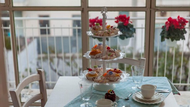 A three-tiered dessert stand on a table with a light blue tablecloth, set against a window with a view of buildings and flowers. - PalazzinApulia (Fasano)