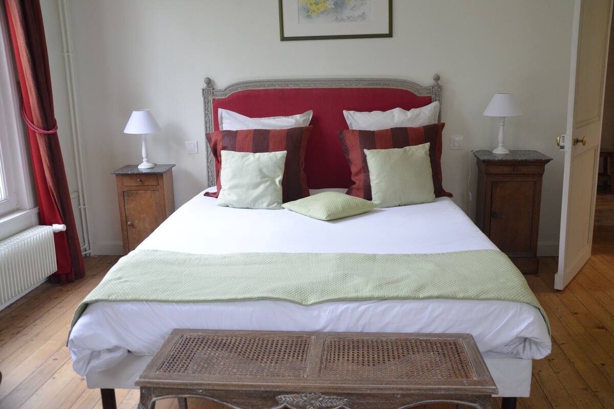 A neatly made bed with a red headboard, two bedside tables with lamps, and a window with curtains.