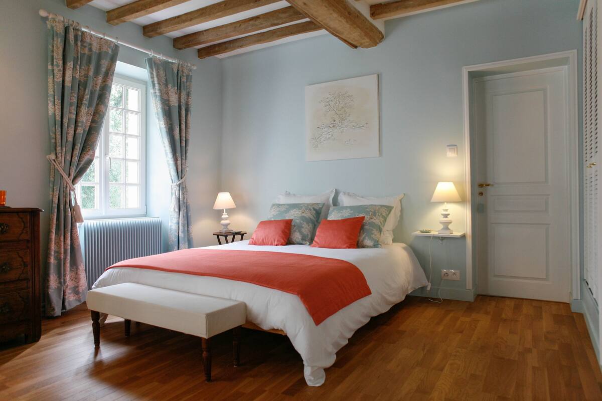 A bedroom with a large bed, wooden floor, a dresser, two bedside tables with lamps, a radiator, and a door.