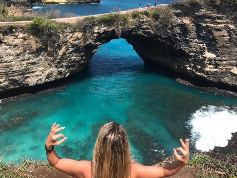 A person sitting on a dirt path with a view of a natural rock arch over a body of water.