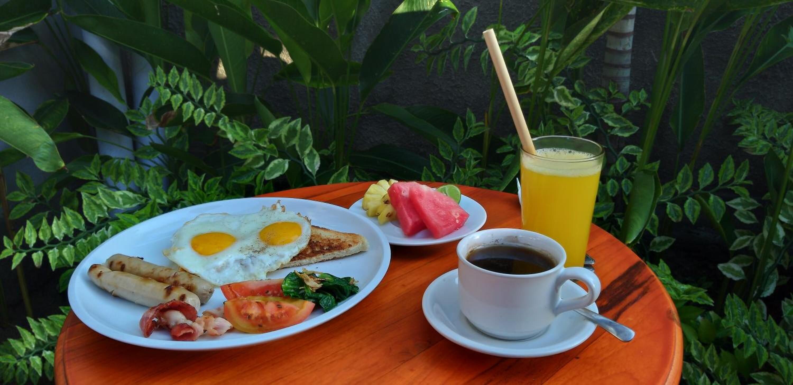 A breakfast plate with eggs, sausages, bacon, tomato, and greens, a cup of coffee, and a glass of orange juice on a wooden table outdoors.