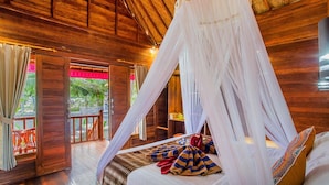 A wooden room with a bed under a canopy, a wooden door, and a view of greenery outside.
