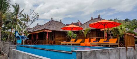 A swimming pool with lounge chairs and umbrellas, surrounded by wooden buildings with tiled roofs.