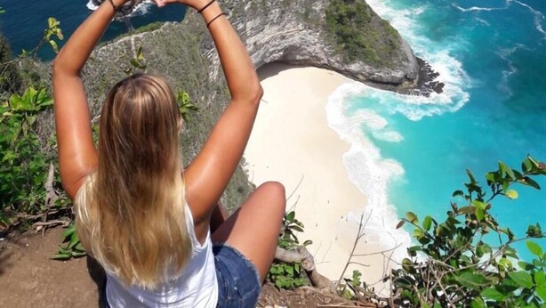 A person sitting on a cliff overlooking a beach with clear blue waters and rocky formations.