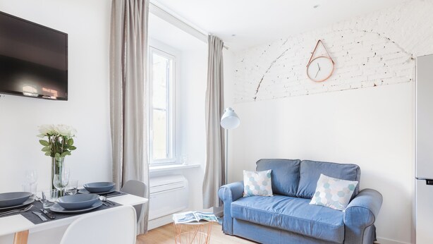 A modern living room with a blue sofa, a dining table set with plates, glasses, and cutlery, a white TV mounted on the wall, and a white brick wall with a clock. - Italianway   - Passeroni 2 (Milan)
